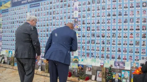 Ukraine Foreign Minister Andrii Sybiha‎ in black suit and his Ghanaian counterpart Samuel Okudzeto Ablakwa in blue suit bow their heads in respect before a memorial wall honouring those killed in the war, as snow falls gently around them