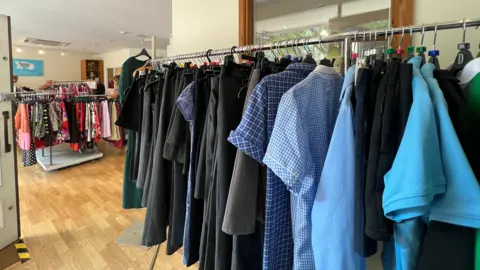 A clothes rack is full of blue tops and shirts and dark grey or black trousers and skirts, with a window behind.
There is another rack of clothes in the background.