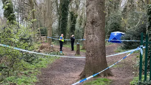 A police cordon attached to series of trees in the middle of a wooded area. A blue police tent can be seen near a footpath on the right hand side of the image. On the left hand side, two police officers are standing and talking to one another.