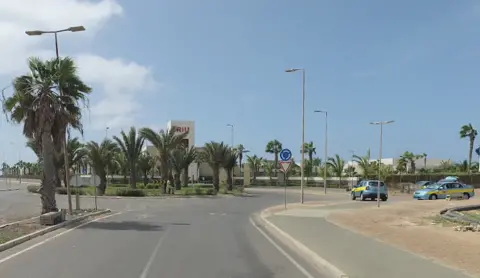Google A road leading up to a roundabout, which has trees in the middle. A hotel is behind the roundabout.