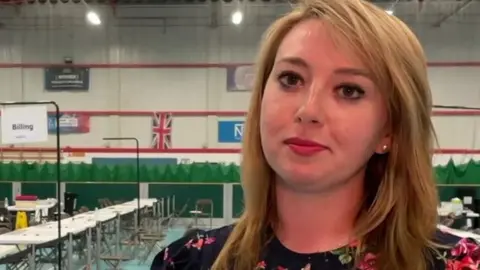Sarah Bool has fair shoulder-length hair and is wearing a black, red and green floral dress. She has a pensive expression and is standing in front of empty tables and chairs at the election cunt in some kind of sports hall.