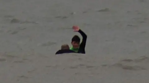 Neil Phillips Oscar is in a grey sea signalling with his arm over his head and the head and shoulders of a man is visible in the water next to him.