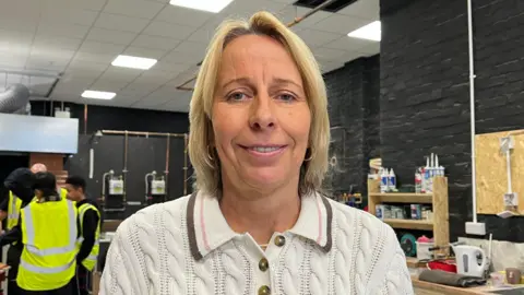 A woman with blonde hair, wearing a light-coloured knitted top. There is a group of young people wearing high visibility jackets behind. There is a black wall with wooden shelving behind her.