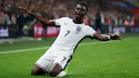 England football player Bukayo Saka knee-sliding across the grass on a football pitch after scoring a goal.
