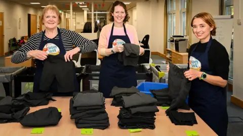 Maraya Gibbs Photography Three workers at Stripey Stork folding uniform items to be administered to children in Surrey. The three women are all wearing navy aprons with the charity's logo, and are all stood beside piles of grey uniform trousers. The lady on the left has blondish hair and is wearing a striped top. The lady in the middle has brown hair and is wearing a pink top. The lady on the right has light brown hair and is wearing a black jumper.