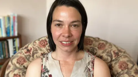 Stef Benstead with shoulder length brown hair and brown eyes is sitting on a chair with a floral pattern wearing a beige top and smiling. 