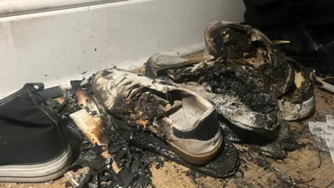 Nicola Rees/ BBC A row of burnt and blackened trainers on the floor is surrounded by ash and other bits of debris.