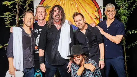 The Foo Fighters band members, with lead singer Dave Grohl in the centre with a towel on his shoulder and his hair wet from sweat, smile at the camera