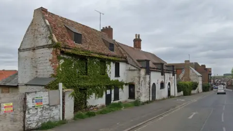 A derelict three-storey building next to a road in Newmarket. It is overgrown with vegetation and weeds and looks in a poor condition.