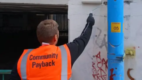 Swindon Borough Council A man in an orange high vis jacket which says 'community payback' paints over some graffiti on a white wall