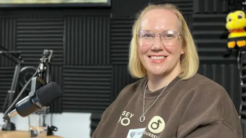 Helen sits at a studio desk in front of a microphone, wearing a brown sweatshirt with printed logos and a lanyard. Behind them are black acoustic foam panels and two plush bee toys mounted on the wall. A framed picture hangs above the microphone setup.