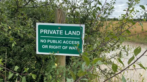 Emily Unia/BBC A hedgerow with a green and white sign saying "Private Land, no public access or right of way" fixed to a wooden post