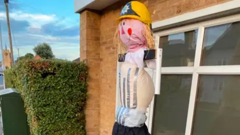 John Devine/BBC A scarecrow of a fireman with its arms and jacket missing. The head and face are still intact with a yellow helmet on its head. Behind is a window and a brick wall with a hedge to the left side of the picture.