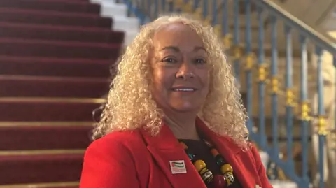 BBC/Claire Hamilton A woman with curly blonde hair, a chunky beaded necklace and a red jacket smiles at the camera whilst standing on a grand staircase
