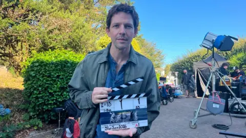 Actor Damien Molony stood on set looking straight at the camera with a Bergerac clapperboard in his hands. There are crews, cameras, lights and filming equipment behind him. 