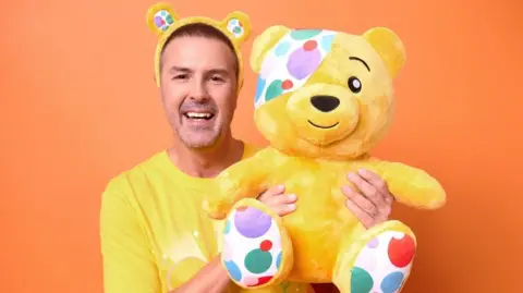 PA Media A man wearing a yellow tshirt and bear ears, holding a yellow bear with a spotted eye patch. He is smiling standing in front of an orange background.