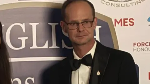 An older man in dinner suit and black tie wearing glasses in front of a backdrop