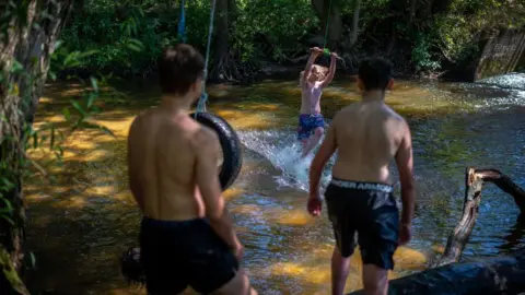 Three youths in shorts play on a rope swing over a river.