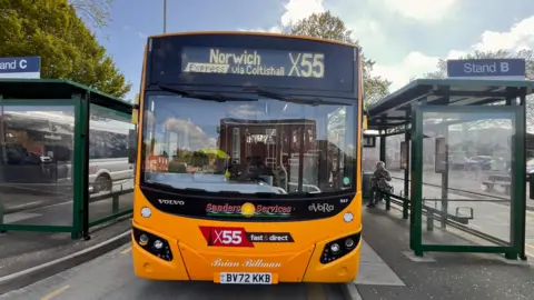 The front of a yellow bus. In the sign at the top it says 'Norwich express via Coltishall X55'. Either side of the bus are bus shelters.