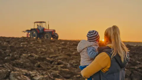 A woman with blonde hair in a grey gillet and long sleeved yellow top. She is holding a toddler in her arms. Both are looking out to a tractor working in a field as the sun sets.