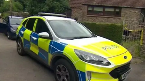 A police car outside the home where Robert Moore was found dead. The car has the usual high-vis blue and yellow markings, and a house built of dark brown bricks can be seen in the background behind a hedge and a curved iron gate with yellow police tape across it. 