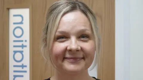 Jamie Niblock/BBC A woman smiles at the camera. She has blonde hair with bangs that frame her face. She is standing in front of a wooden door.