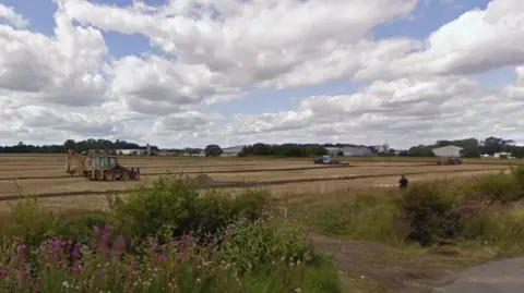 Google A field which is the site of the proposed solar farm. Three farm vehicles are in the field.