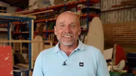 A bald man smiles at the camera, and he is wearing a pale blue polo shirt. In the background there are pieces of wood to build a float for the Battle of Flowers parade.