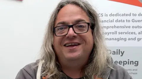 BBC A man with long grey hair and black glasses is wearing a grey hoodie and T shirt he is in front of a TCS poster