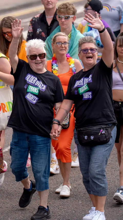 PA Media Two people wearing matching black T-shirts reading “Older Dykes for Trans Rights” walk hand in hand at a Pride parade. Both wave toward the camera as a crowd of participants follows behind them along a city street.