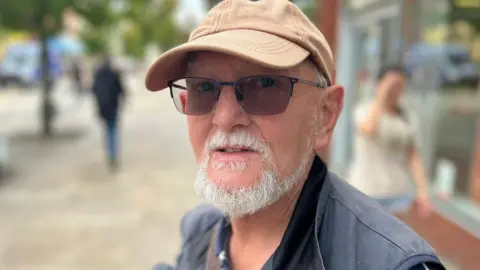 Keith Andrews, with a white beard, wears sunglasses, a beige cap and a navy jacket. 