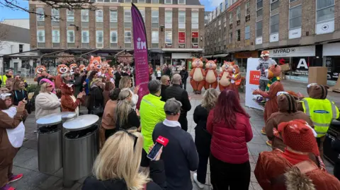 A large group of people in different squirrel costumes are looking at another man on standing on a bench who has a mega phone speaking to the crowd.