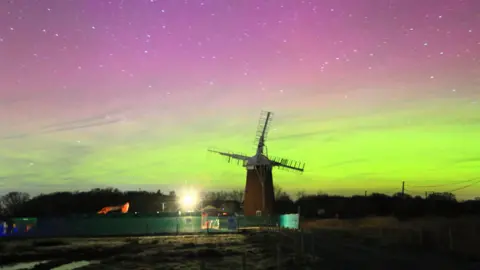 BBC Weather Watchers/KeepOnRunning A windmill is in the middle and has a green fence around it. There are trees in the distance and the Northern Lights have made the sky turn purple, pink, and yellow, and there are several stars.