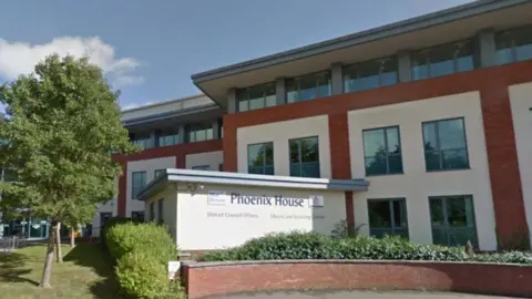 A building with brown brickwork and several windows stands before a sign which says Phoenix House, under which is written District Council Offices. 