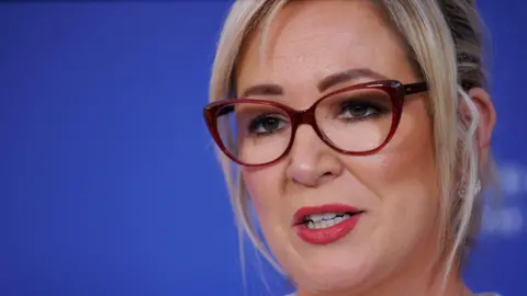 A woman with blonde hair looks to the left of the camera. She is wearing glasses. Behind her is a blue background. 