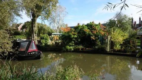 A black narrowboat, with a red triangular front, is coming into the picture - travelling down the canal. There are trees and bushes with their leaves just starting to turn orange and brown on the far bank