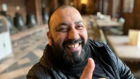 A headshot of a man sitting in a cathedral. He is smiling at the camera and has his thumb up. He is bald and has a black beard. 