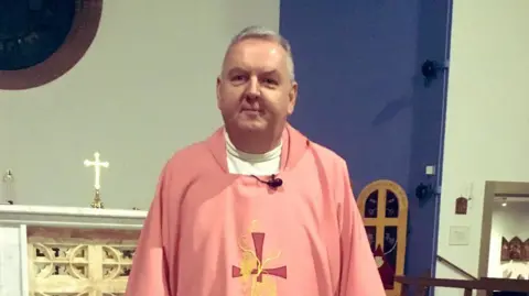 A priest, who is an older man with short grey hair, is smiling and standing at the front of a Catholic church. He is wearing a pink robe which has a cross on it. 