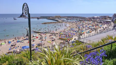 Love Lyme Regis A beach with hundreds of people on. There are lots of parasols and windbreakers. The photo is taken from a hill looking down. In the foreground there is a lamp post with an ammonite shape on it.