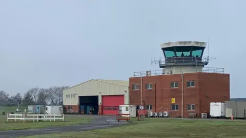 A view of the control tower at Coventry Airport showing a glass hexagonal tower on top of a red brick building, which is next to a big metal hanger.