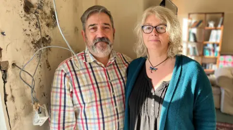 BBC Tony and Becs Wadley are standing in the living room of their Gower Peninsula home. On the wall next to them there is black mould and an exposed electrical socket after internal wall insulation was badly installed.
