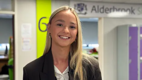 Year 13 student Jasmine has blonde hair and brown eyes. She wears a black blazer and white shirt and is smiling. She is standing in the sixth form corridor with some of the common rooms behind her.