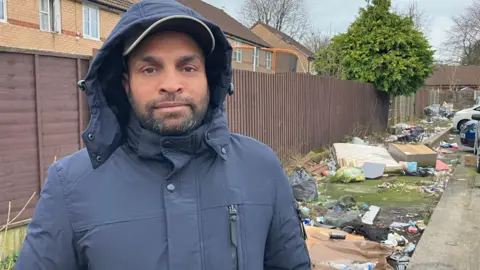 Muhammad Amin Malik is looking into the camera and is wearing a blue rain jacket and black cap. Behind him is piles of rubbish, including a mattress, old wardrobe and black bags. 