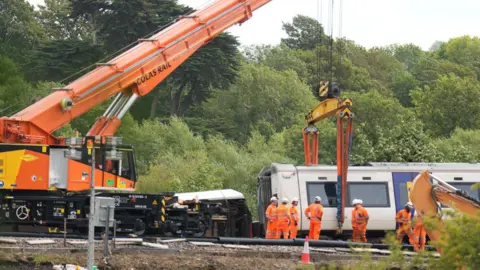 A large orange rail crane towers above a Southern train carriage as recovery crews prepare it for lifting. Several workers in orange high-visibility clothing stand beside the carriage on the railway line, with trees and vegetation surrounding the site.