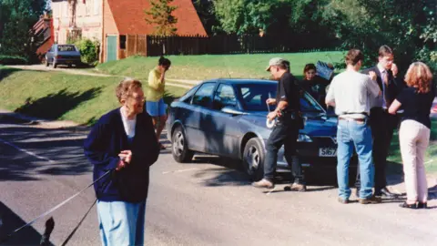 Carol Sawbridge A woman walking her dogs past the crew filing Midsomer Murders in Ewelme in the 1990s. She is smiling. Several crew members are gathered around a car, some are talking on the phone.