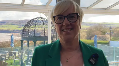 Beverley Rutherford is a woman in her forties wearing a green jacket with a white top and with short blonde hair and glasses. She is standing in the garden of a hotel on decking with several tables and chairs on grass behind with blue umbrellas folded down. The garden overlooks a wide beach. 