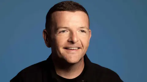Studio-style head and shoulders shot of a smiling Kevin Bridges. he is wearing a black top and has been photographed against a blue backdrop.