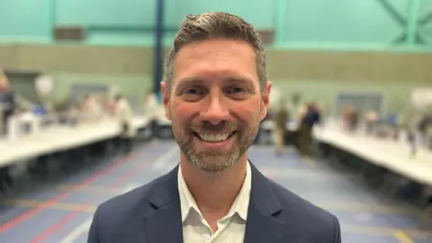 A man from torso up wearing a suit with brown and grey hair and grey stubble. A sportshall is out of focus in the background.