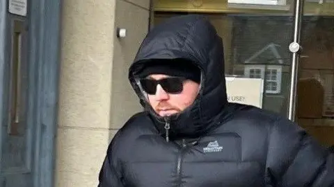 David Gowans wearing a black padded jacket with the hood up. He is also wearing a black hat and sunglasses as he leaves court.