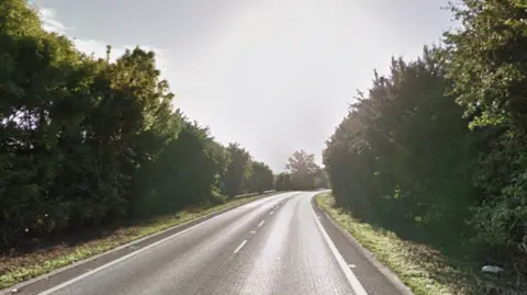 An empty stretch of A-road. There are trees on either side of the road and bright sun. 